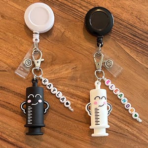 May include: Two retractable badge holders with syringe charms. One is black with the name "KAYLA" and the other is white with the name "ELEANOR". The syringe charms have a smiling face and measurement markings. The badge holders are white and black.