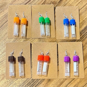 Puede incluir: Una colección de seis pares de pendientes originales, cada uno con un pequeño tubo de plástico transparente y una tapa de color. Las tapas son de color naranja, verde, azul, marrón, rojo y morado. Cada pendiente está unido a una pequeña tarjeta marrón.