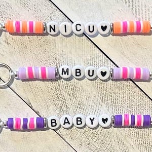 Puede incluir: Tres llaveros de cuentas con cierres plateados. Cada llavero tiene una palabra diferente escrita con cuentas de letras blancas: NICU, MBU y BABY. Los llaveros presentan cuentas a rayas de colores en tonos de rosa, morado y naranja, con cuentas en forma de coraz&oacute;n.