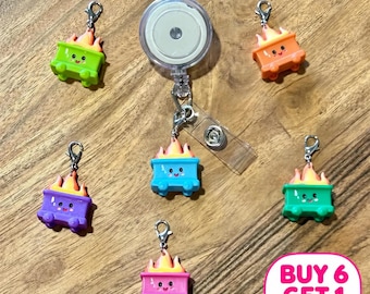 May include: A retractable badge reel with a clear plastic strap and a metal snap, accompanied by six charms shaped like miniature dumpsters on fire. Each dumpster features a smiling face and comes in green, orange, blue, purple, teal, or pink.