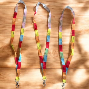 Puede incluir: Tres cordones multicolores con un patrón repetitivo de bloques rectangulares. Cada cordón tiene un cierre plateado. Los cordones están sobre una superficie de madera de color marrón claro.