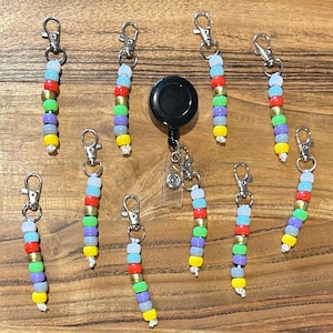 May include: A selection of beaded keychains and a retractable badge holder. The keychains have colourful beads in a rainbow pattern, including yellow, purple, green, red, and blue, with silver-tone metal clasps. The badge holder is black.