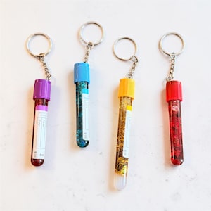 Puede incluir: Cuatro llaveros cilíndricos que parecen tubos de ensayo de laboratorio de 5 ml con tapas de colores morado, azul, amarillo y rojo. Cada tubo contiene purpurina a juego y una etiqueta blanca con texto impreso y líneas para anotar datos.