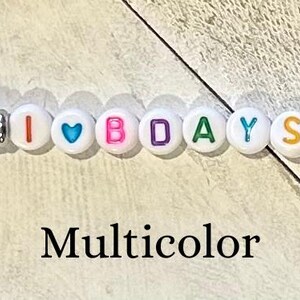 Puede incluir: Un llavero con un cierre plateado y un dise&ntilde;o de cuentas. Las cuentas son blancas con las letras "I ♡ BDAYS" y acentos coloridos de rosa, azul y blanco. La palabra "Multicolor" est&aacute; debajo.
