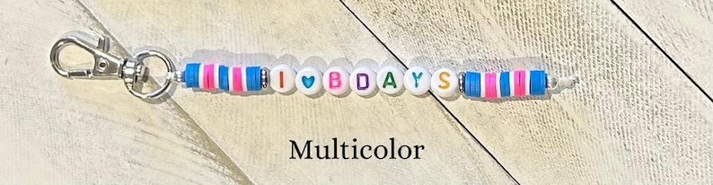 Puede incluir: Un llavero con un cierre plateado y un dise&ntilde;o de cuentas. Las cuentas son blancas con las letras "I ♡ BDAYS" y acentos coloridos de rosa, azul y blanco. La palabra "Multicolor" est&aacute; debajo.