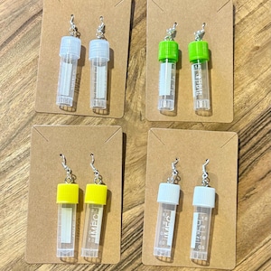 Puede incluir: Cuatro pares de pendientes novedosos, cada uno con un vial de pl&aacute;stico transparente en miniatura con una tapa de color. Las tapas son blancas, verdes y amarillas. Los viales est&aacute;n unidos a ganchos para las orejas plateados y se muestran en peque&ntilde;as tarjetas marrones.
