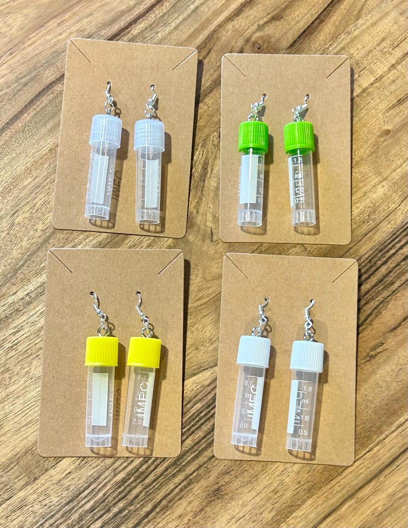 Puede incluir: Cuatro pares de pendientes novedosos, cada uno con un vial de pl&aacute;stico transparente en miniatura con una tapa de color. Las tapas son blancas, verdes y amarillas. Los viales est&aacute;n unidos a ganchos para las orejas plateados y se muestran en peque&ntilde;as tarjetas marrones.
