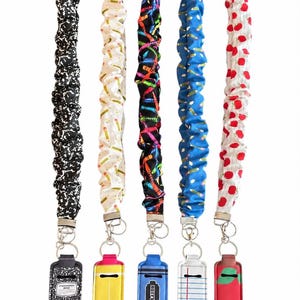 May include: Five fabric lanyards with matching rectangular holders. Patterns include black and white composition book, yellow pencils on white, colorful crayons on black, blue with pencils, and red apples on white pinstripes with metal key rings.