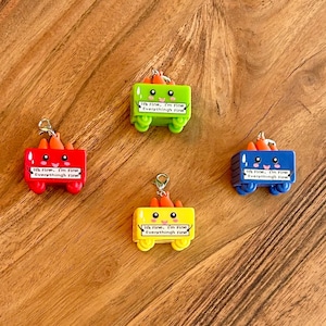 Może przedstawiać: Cztery kolorowe, miniaturowe zawieszki w kształcie wagon&oacute;w z pomarańczowymi akcentami przypominającymi marchewki. Każdy wagon jest w innym kolorze: czerwonym, zielonym, ż&oacute;łtym i niebieskim. Tekst "It's Fine, I'm Fine, Everything's Fine" jest wydrukowany na każdym wagonie.