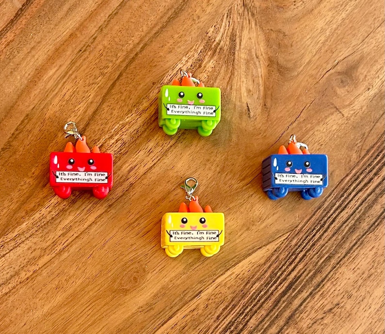 Może przedstawiać: Cztery kolorowe, miniaturowe zawieszki w kształcie wagon&oacute;w z pomarańczowymi akcentami przypominającymi marchewki. Każdy wagon jest w innym kolorze: czerwonym, zielonym, ż&oacute;łtym i niebieskim. Tekst "It's Fine, I'm Fine, Everything's Fine" jest wydrukowany na każdym wagonie.