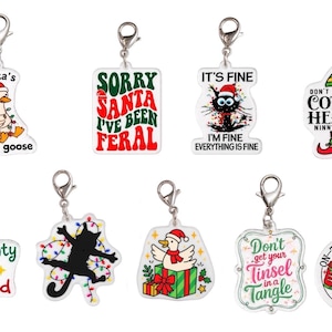 Puede incluir: Un conjunto de nueve dijes de acrílico con cierres de langosta plateados, que presentan ilustraciones y textos con temática navideña, incluyendo Santa's silliest goose, Sorry Santa I've been feral, It's fine, Cotton headed ninny muggins, Naughty Nice I Tried.