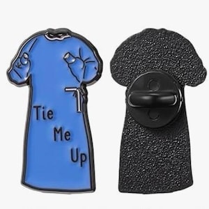 Könnte beinhalten: Ein blauer Emaille-Pin in Form eines OP-Kittels mit den Worten "Tie Me Up" in Weiß. Der Pin hat eine schwarze Rückseite mit einem Butterfly-Verschluss. Das Design des Kittels umfasst Ärmel und ein Bindedetail.