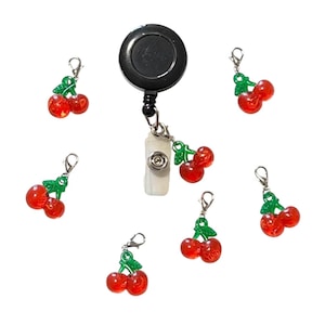 May include: A black retractable badge reel with a white plastic clip and several red cherry charms with green leaves. The charms are attached with silver-colored clasps.