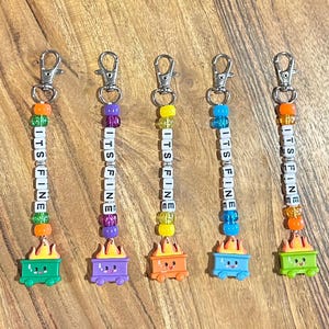 May include: Five keychains with silver clasps. Each keychain has a beaded strand with the words "ITS FINE" spelled out in white letter beads. The keychains are topped with a colorful, cartoonish wagon with flames.