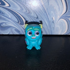 Puede incluir: Un pequeño juguete monstruo azul y verde con grandes ojos brillantes. El juguete tiene una sonrisa de dientes y está sentado sobre una superficie oscura.