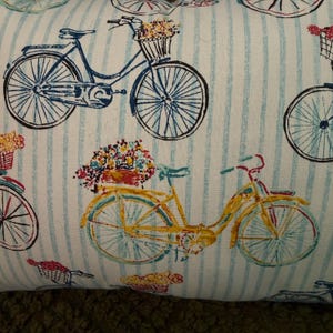May include: A fabric cushion cover featuring a repeating pattern of bicycles on a white background with vertical light blue stripes. The bicycles are depicted in blue and yellow, each equipped with a front basket filled with colourful flowers.