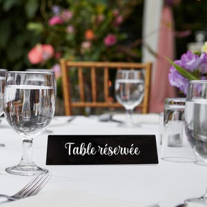 Peut inclure: Une table de réception avec des verres à eau transparents, des couverts et un panneau noir avec l'inscription "Table réservée" en écriture blanche. La table est recouverte d'une nappe blanche, avec un arrière-plan flou de verdure et de fleurs.