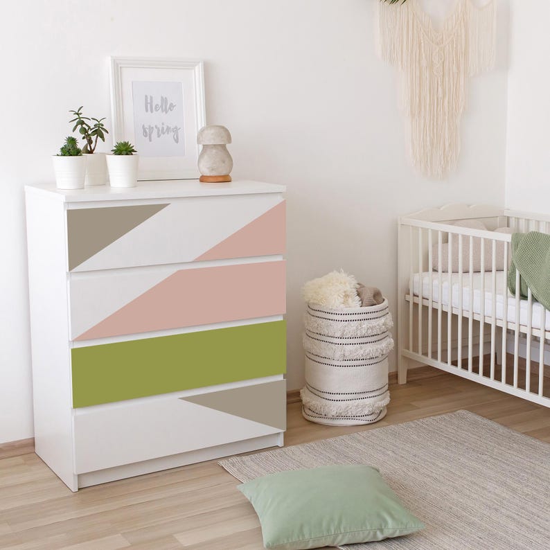 K&ouml;nnte beinhalten: Wei&szlig;e Kommode mit vier Schubladen, jede mit einem geometrischen Design in den Farbt&ouml;nen Taupe, Rosa und Gr&uuml;n. Ein gerahmter Druck mit dem Text "Hello spring" befindet sich oben, zusammen mit kleinen Topfpflanzen und einem dekorativen Pilz.