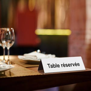 Peut inclure: Une pancarte de table blanche avec le texte "Table réservée" en noir est posée sur une table en bois sombre. Des verres à vin et une serviette pliée sont en arrière-plan, suggérant un restaurant.