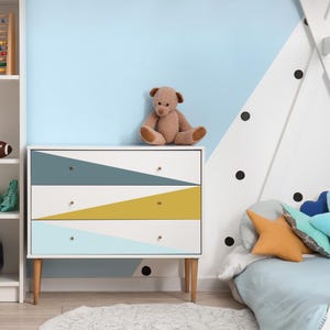K&ouml;nnte beinhalten: Eine wei&szlig;e Kommode mit drei Schubladen und geometrischen Designs in T&uuml;rkis, Gelb und Hellblau. Sie hat Holzbeine und Messingkn&ouml;pfe. Ein brauner Teddyb&auml;r sitzt oben drauf. Der Raum hat eine hellblaue und wei&szlig;e Wand.