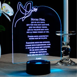 Könnte beinhalten: Ein blaues LED-Licht mit einer weißen Umrandung einer Taube und eines Zweigs. Das Licht zeigt das Vaterunser auf Französisch, "NOTRE PÈRE, QUI ES AUX CIEUX, QUE TON NOM SOIT SANCTIFIÉ, QUE TON RÈGNE VIENNE, QUE TA VOLONTÉ SOIT FAITE SUR LA TERRE COMME AU CIEL. DONNE-NOUS AUJOURD'HUI NOTRE PAIN DE CE JOUR. PARDONNE NOUS NOS OFFENSES, COMME NOUS PARDONNONS AUSSI À CEUX QUI NOUS ONT OFFENSÉS. ET NE NOUS LAISSE PAS ENTRER EN TENTATION, MAIS DÉLIVRE-NOUS DU MAL. AMEN."