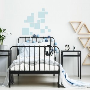K&ouml;nnte beinhalten: Ein schwarzes Metallbett mit wei&szlig;er Bettw&auml;sche, blauen Kissen und einer grauen Decke. Das Bett ist mit blau-wei&szlig;en Lichterketten geschm&uuml;ckt. Ein Kopfh&ouml;rer h&auml;ngt am Rahmen. Ein schwarzer Nachttisch mit einer Uhr und einer Tasse.