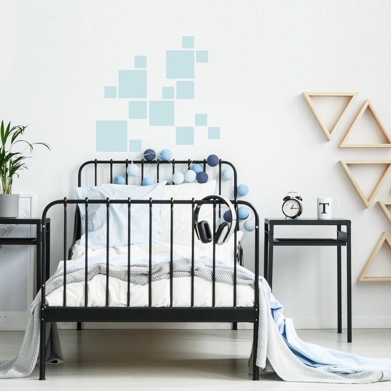 K&ouml;nnte beinhalten: Ein schwarzes Metallbett mit wei&szlig;er Bettw&auml;sche, blauen Kissen und einer grauen Decke. Das Bett ist mit blau-wei&szlig;en Lichterketten geschm&uuml;ckt. Ein Kopfh&ouml;rer h&auml;ngt am Rahmen. Ein schwarzer Nachttisch mit einer Uhr und einer Tasse.