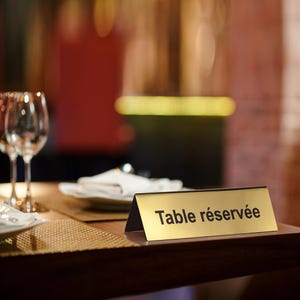 Peut inclure: Un panneau de table doré avec les mots "Table réservée" en texte noir. Le panneau est posé sur une table en bois sombre, avec des verres à vin et une serviette pliée visibles en arrière-plan. Le cadre suggère un restaurant ou un événement.