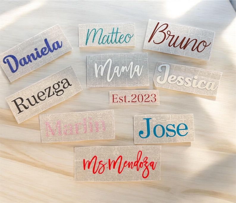 May include: A collection of personalized name decals in various colors and fonts. Names include "Daniela," "Matteo," "Bruno," "Ruezga," "Mama," "Jessica," "Marlin," "Jose," "Ms. Mendoza," and "Est. 2023."