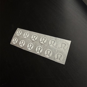May include: A sheet of small, circular stickers with a white logo on a transparent background. The logo is a stylized omega symbol. The stickers are arranged in rows and columns on a rectangular sheet, and are on a black surface.