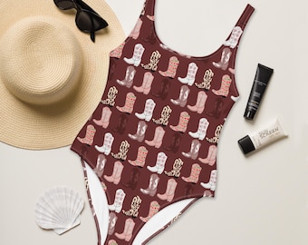Könnte beinhalten: Ein einteiliger Badeanzug mit burgunderfarbenem Hintergrund und einem sich wiederholenden Muster von Cowboy-Stiefeln in verschiedenen Farben und Designs. Der Badeanzug wird mit einem Strohhut, Sonnenbrille, Sonnencreme und einer Muschel gezeigt.