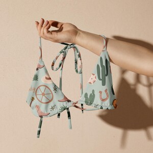 May include: A light blue bikini top with a repeating pattern of cowboy-themed illustrations, including cacti, cowboy boots, and a horseshoe. The top is held up by a hand, showcasing the tie-up straps and triangle shape.