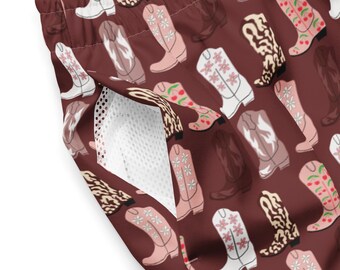 May include: Brown swim trunks with a repeating pattern of cowboy boots in various colors and designs. The boots include floral, cherry, and patterned accents. The trunks have a white mesh pocket.