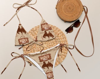Könnte beinhalten: Ein brauner und beiger Bikini mit weißem Hintergrund. Das Bikinioberteil und -unterteil haben ein braunes Motiv eines Cowboys, der ein Pferd reitet, mit dem Text "Rodeo Vibes". Der Bikini liegt auf einer geflochtenen Strohhütte mit einer braunen geflochtenen Tasche und einer Sonnenbrille.