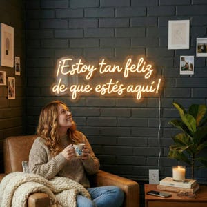 Puede incluir: Una mujer sentada en un sillón de cuero marrón, sosteniendo una taza, debajo de un letrero de neón que dice "¡Estoy tan feliz de que estés aquí!" en amarillo cálido. La habitación tiene una pared de ladrillo oscuro, con fotos enmarcadas y una mesa auxiliar.