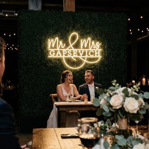 Może przedstawiać: Scena przyjęcia weselnego z neonowym napisem "Mr & Mrs Gapsevich" nad parą śmiejącą się. Na stole znajduje się kompozycja kwiatowa i kieliszki do wina. Tło to zielona ściana.