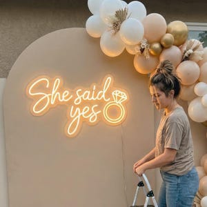 May include: A neon sign that reads "She said yes" with a ring graphic, set against a beige backdrop. The sign is part of a decorative display with balloons in white, gold, and beige tones, suggesting a celebration or event.