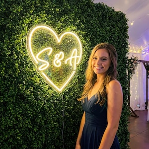 Op de afbeelding: Een neonbord in de vorm van een hart met de letters "S & A" in een sierlijk lettertype. Het bord is verlicht met een warme, gele gloed en staat tegen een achtergrond van groen gebladerte. Een vrouw in een marineblauwe jurk glimlacht.