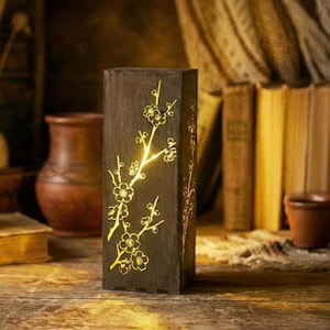 May include: A dark brown wooden lantern with a floral design. The lantern is rectangular and features cut-out floral patterns that illuminate with a warm, yellow light. The lantern is placed on a wooden surface.