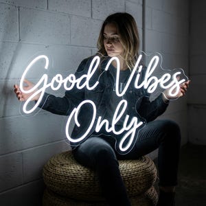 May include: A white neon sign with the words "Good Vibes Only" in a cursive font. The sign is held by a person wearing a denim jacket and dark jeans. The background is a light gray brick wall.
