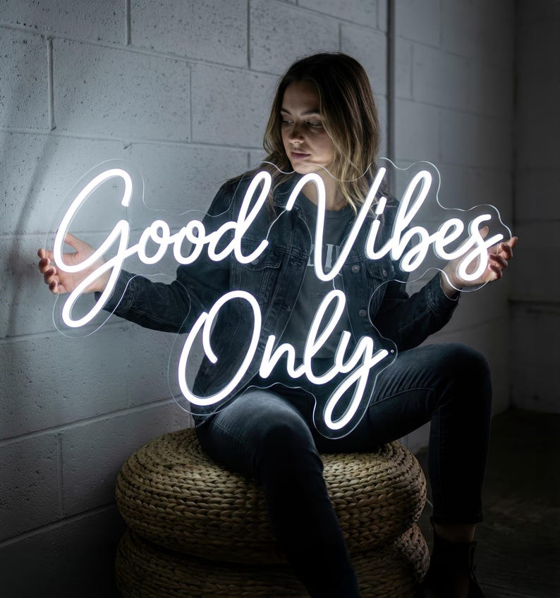 May include: A white neon sign with the words "Good Vibes Only" in a cursive font. The sign is held by a person wearing a denim jacket and dark jeans. The background is a light gray brick wall.