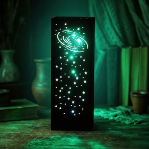 May include: A black rectangular night light with a galaxy design. The light features a glowing white galaxy and numerous white stars. The light emits a soft green glow, creating a calming ambiance. The night light is placed on a wooden surface.