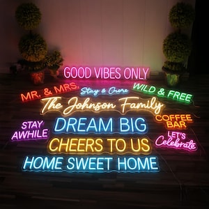 May include: A collection of neon signs in various colors, including pink, yellow, blue, and green. The signs display phrases such as "Good Vibes Only," "Dream Big," and "Home Sweet Home." The signs are arranged on a dark surface.