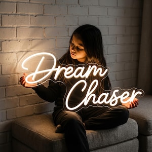 Peut inclure: Une enseigne au néon blanche en écriture cursive avec l'inscription "Dream Chaser". L'enseigne est tenue par une personne devant un mur de briques. L'enseigne est éclairée et brille vivement.