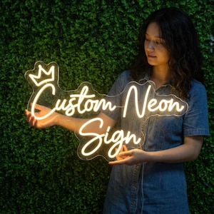 May include: A neon sign in the shape of a crown, with the words "Custom Neon Sign" in a cursive font. The sign is held by a person against a green, leafy backdrop. The sign glows with a warm, inviting light.