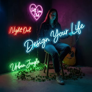 May include: A woman sits with a neon sign that says "Design Your Life" in cursive. Other neon signs include a heart that says "Self Love Club", "Night Owl", and "Urban Jungle". A guitar is in the background.