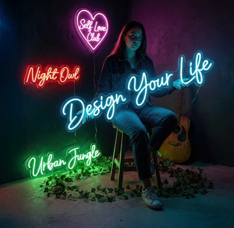 May include: A woman sits with a neon sign that says "Design Your Life" in cursive. Other neon signs include a heart that says "Self Love Club", "Night Owl", and "Urban Jungle". A guitar is in the background.