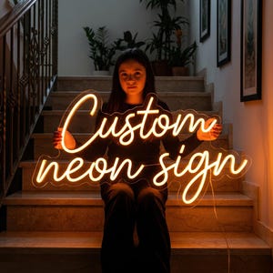 May include: A warm, glowing neon sign with the words "Custom neon sign" in a cursive font. The sign is held by a person sitting on a staircase. The sign emits a soft, inviting light.