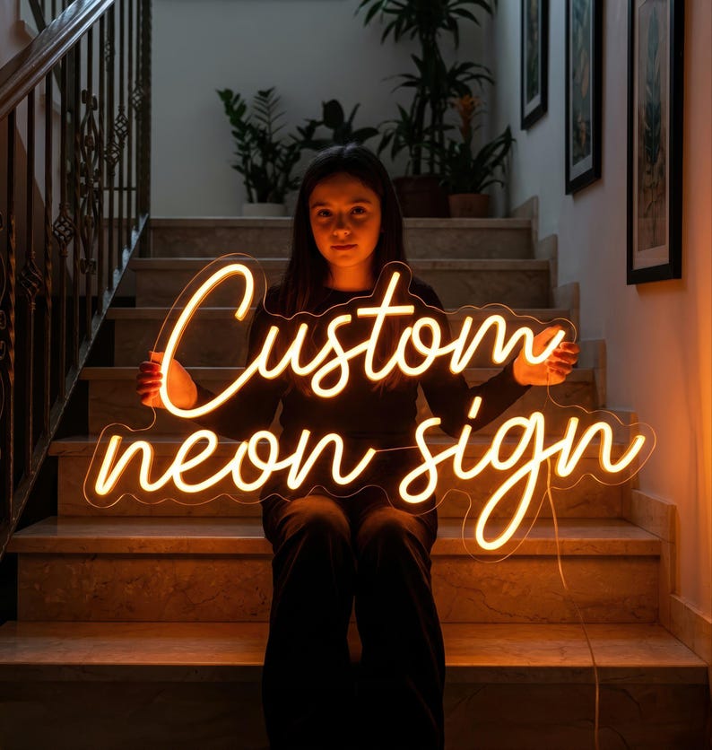 May include: A warm, glowing neon sign with the words "Custom neon sign" in a cursive font. The sign is held by a person sitting on a staircase. The sign emits a soft, inviting light.