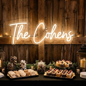 May include: A glowing neon sign that reads "The Cohens" hangs above a table laden with treats. The table features cupcakes, cookies, and jars of candies, all set against a rustic wooden backdrop, creating a warm, inviting atmosphere.